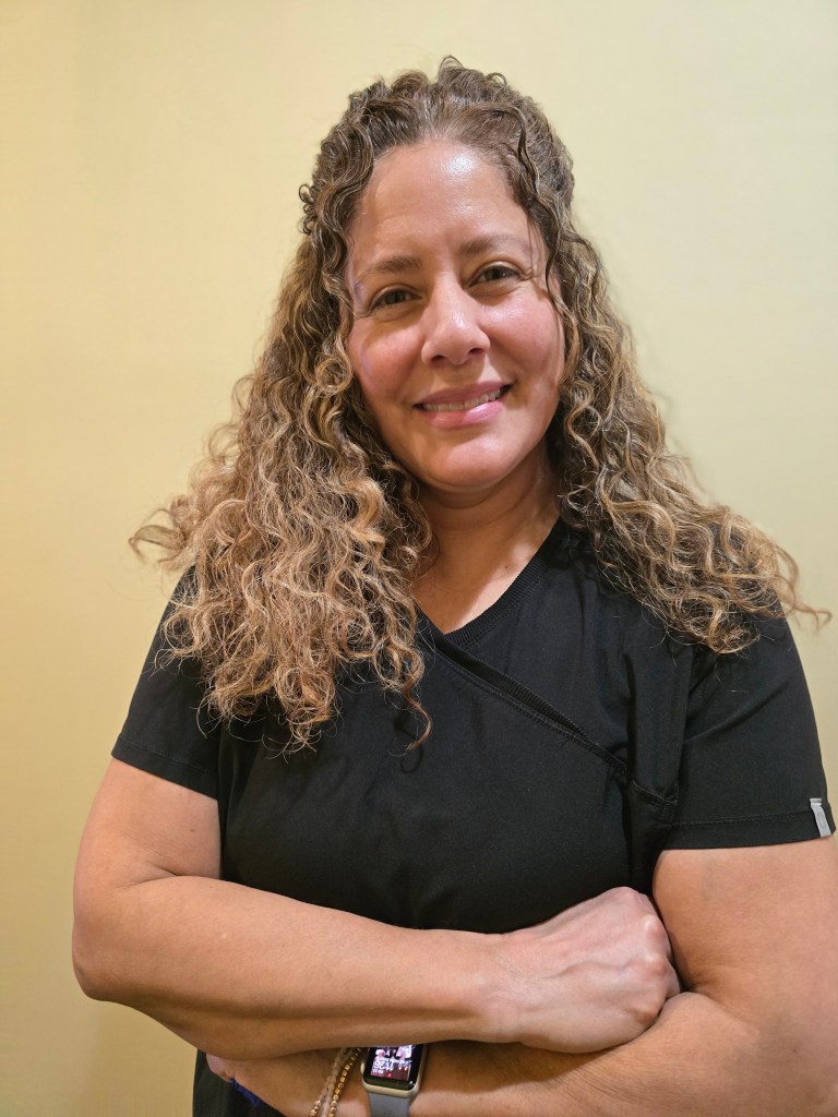 A woman with curly hair stands confidently, smiling with her arms crossed. She is wearing a black shirt and has a watch on her wrist, with a soft yellow background.
